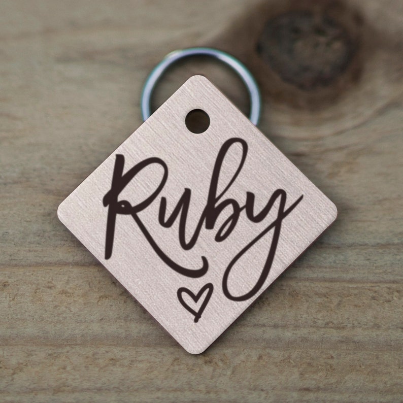 May include: A silver diamond-shaped dog tag with a black cursive inscription that reads "Ruby" and a small heart symbol.