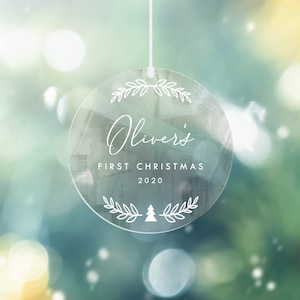 K&ouml;nnte beinhalten: Ein runder, transparenter Weihnachtsbaumschmuck mit einer wei&szlig;en Schnur zum Aufh&auml;ngen. Der Schmuck hat ein wei&szlig;es Design mit dem Text "Oliver's First Christmas 2020" und einem kleinen Weihnachtsbaum.