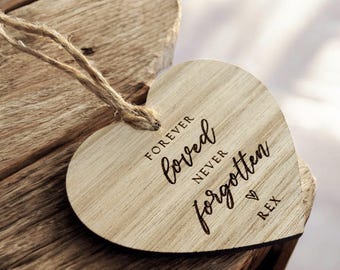 May include: A wooden heart-shaped ornament with the words "Forever Loved Never Forgotten" engraved in a script font. A small heart and the name "REX" are also engraved. The ornament has a jute twine loop for hanging.