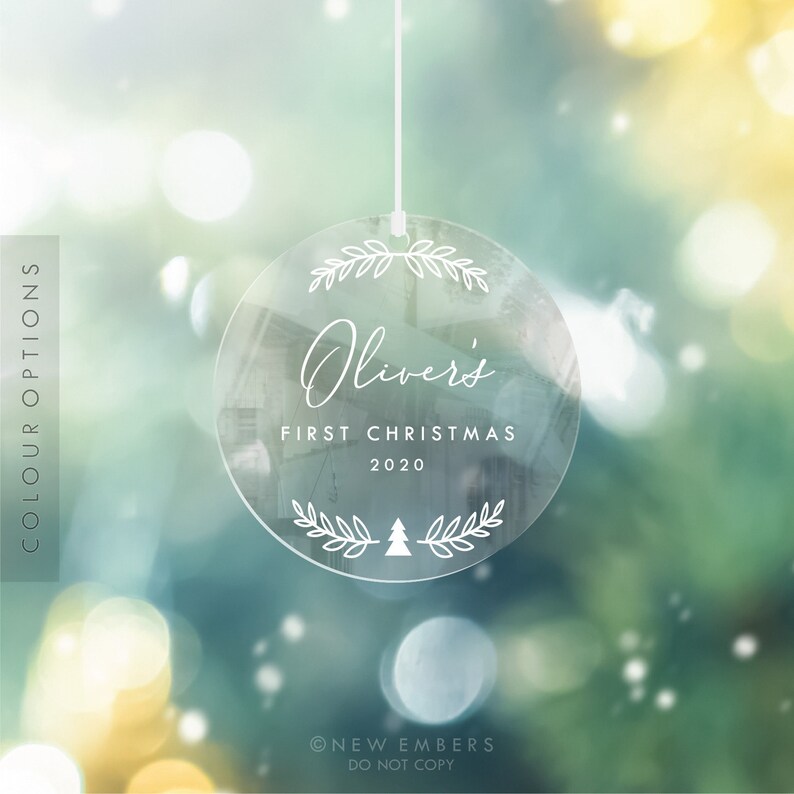 K&ouml;nnte beinhalten: Ein runder, transparenter Weihnachtsbaumschmuck mit einer wei&szlig;en Schnur zum Aufh&auml;ngen. Der Schmuck hat ein wei&szlig;es Design mit dem Text "Oliver's First Christmas 2020" und einem kleinen Weihnachtsbaum.