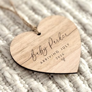 May include: A heart-shaped wooden ornament with the text "Baby Parker ARRIVING JULY 2026" engraved in a delicate script. A small heart symbol is below the date. The ornament hangs from a natural twine string.