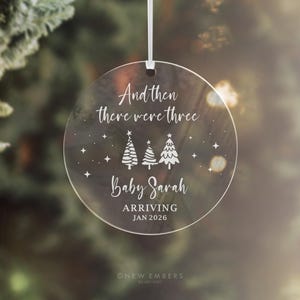 May include: Clear, round Christmas ornament with white text and a white ribbon. The text reads "And then there were three" above three Christmas trees and the name "Baby Sarah" with the arrival date "JAN 2026".