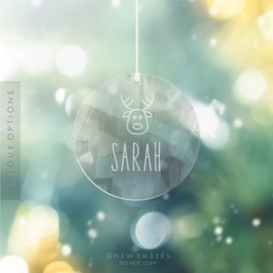 May include: A clear glass bauble with a white reindeer and the name "SARAH" printed on it. The bauble is hanging from a white string against a blurred background of green and yellow bokeh.