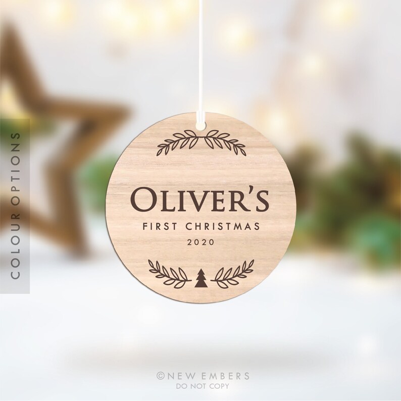 May include: A round wooden ornament with a brown woodgrain finish. The ornament features the text "Oliver's First Christmas 2020" with a small Christmas tree graphic and a leafy wreath design.
