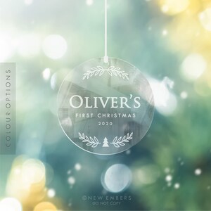 May include: A clear round Christmas ornament with a white string hanging from the top. The ornament has a white design of a wreath, the name "Oliver's", the text "First Christmas 2020", and a small Christmas tree inside the wreath.