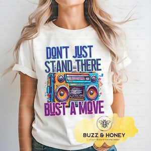 以下が含まれることがあります： クリーム色のTシャツ。カラフルなレトロなラジカセのグラフィックが特徴です。「DON'T JUST STAND THERE」の文字がラジカセの上に、「BUST A MOVE」の文字が下にあります。半袖でクルーネックのシャツです。