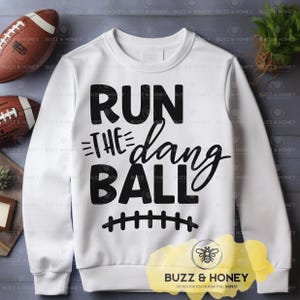 May include: White sweatshirt featuring the phrase "RUN THE dang BALL" in black, with a football graphic. A crew neck style, this sweatshirt is ideal for football enthusiasts. The text is in a bold font.