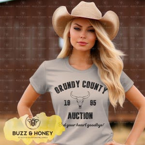 以下が含まれることがあります： 「Grundy County Auction」の文字と雄牛の頭蓋骨のグラフィックがプリントされたライトグレーのTシャツ。シャツには「1995」の年号と「Bid your heart goodbye!」のフレーズも表示されています。