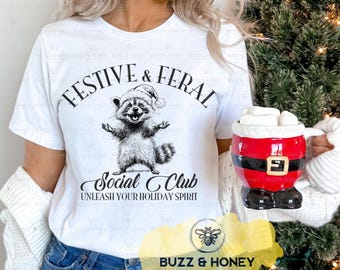 Może przedstawiać: Biała koszulka z napisem "Festive & Feral Social Club" i szopem w czapce Świętego Mikołaja. Czerwono-czarny kubek w tematyce Świętego Mikołaja trzymany jest w prawej ręce. Koszulka ma grafikę szopa z podniesionymi ramionami.