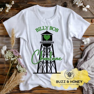 May include: White t-shirt featuring a black and green graphic of a water tower with the names "Billy Bob" and "Charlene" in green. The water tower design includes a green heart. The shirt also has the text "Buzz & Honey".