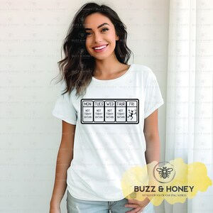 May include: White t-shirt with a black graphic. The graphic features a calendar-like design with the days of the week and the text "NOT TODAY SATAN". The Friday box has a devil illustration. The shirt is a casual wear item.