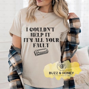 Könnte beinhalten: Ein beiges T-Shirt mit dem Text "I COULDN'T HELP IT IT'S ALL YOUR FAULT" in Schwarz und einer Mundharmonika-Illustration. Das Shirt zeigt auch das Logo "BUZZ & HONEY". Das Shirt ist Teil einer Bekleidungslinie.