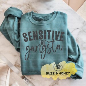 K&ouml;nnte beinhalten: T&uuml;rkisfarbenes Sweatshirt mit dem Text "SENSITIVE lil' gangsta" in dunkelgrauen Buchstaben. Das Sweatshirt liegt gefaltet auf einer Marmoroberfl&auml;che. Das Logo von Buzz & Honey ist unten rechts sichtbar.