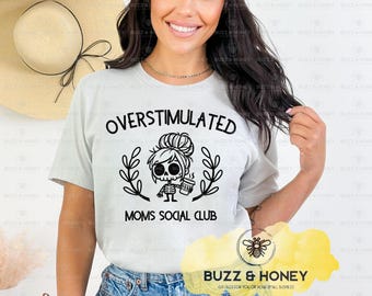 Op de afbeelding: Lichtgrijs t-shirt met de tekst "OVERSTIMULATED" boven een afbeelding van een skeletvrouw met een koffiekopje, omringd door bladeren. Daaronder staat "MOMS SOCIAL CLUB". Op de achtergrond is een grote strohoed te zien.