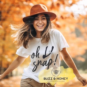 May include: A woman wearing a white t-shirt with the words "oh snap!" in black script. She is also wearing a brown hat. The background is blurred with autumn colours.