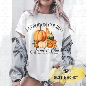 Op de afbeelding: Wit T-shirt met een kleurrijke afbeelding van pompoenen in verschillende maten en tinten oranje, groen en crème. De tekst luidt "FAT BOTTOM GOURDS Social Club" met de slogan "WE MAKE THE ROCKIN' WORLD GO ROUND".