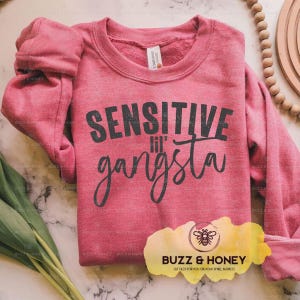 K&ouml;nnte beinhalten: Ein meliertes rotes Sweatshirt mit dem Text "SENSITIVE lil' gangsta" in Schwarz. Das Sweatshirt hat lange &Auml;rmel und einen Rundhalsausschnitt. Das Bild enth&auml;lt auch ein Logo f&uuml;r "Buzz & Honey" und eine Holzperlen-Girlande.