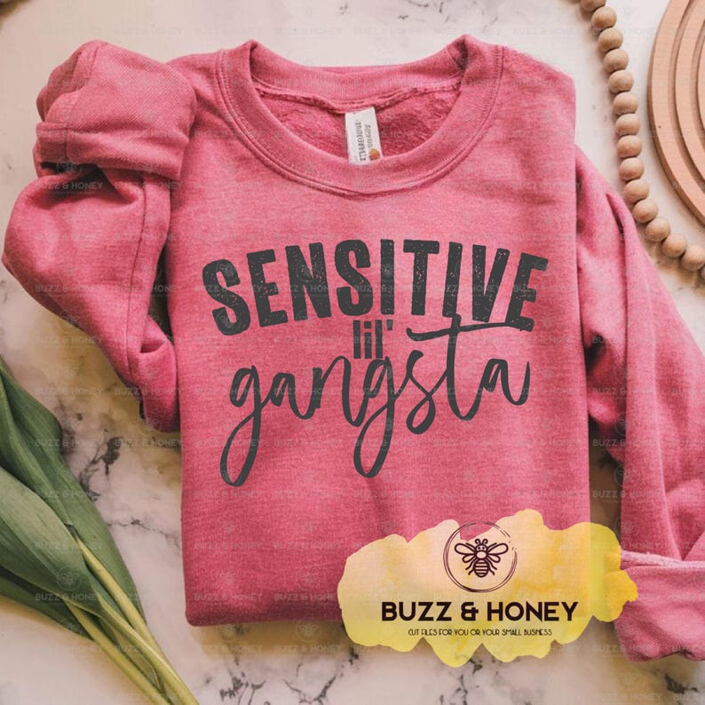 K&ouml;nnte beinhalten: Ein meliertes rotes Sweatshirt mit dem Text "SENSITIVE lil' gangsta" in Schwarz. Das Sweatshirt hat lange &Auml;rmel und einen Rundhalsausschnitt. Das Bild enth&auml;lt auch ein Logo f&uuml;r "Buzz & Honey" und eine Holzperlen-Girlande.