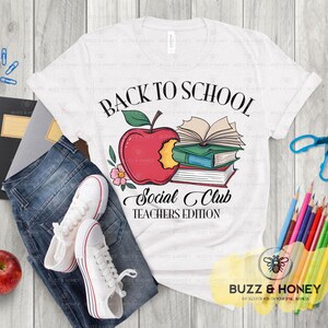 May include: White t-shirt with a "Back to School Social Club Teachers Edition" graphic featuring a red apple, books, and floral accents. The shirt is paired with blue jeans and white sneakers.