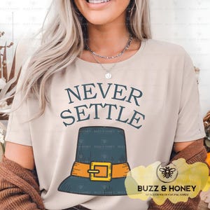 May include: Beige t-shirt with the text "NEVER SETTLE" above a blue pilgrim hat with an orange belt and gold buckle. The shirt has a round neck and short sleeves. The Buzz & Honey logo is at the bottom right.