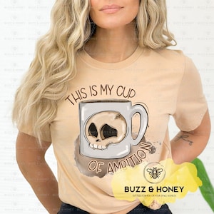 Puede incluir: Una camiseta de color beige con un gráfico de una taza de café con una calavera en su interior. El texto de la camiseta dice "This is my cup of ambition".
