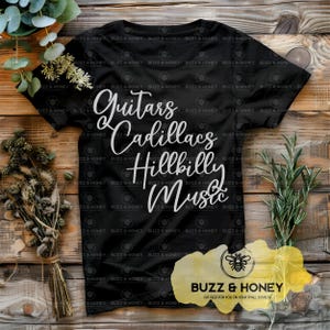 May include: Black t-shirt with the words "Guitars, Cadillacs, Hillbilly Music" in white script. The shirt also has the logo "Buzz & Honey" in the bottom right corner. The shirt is displayed on a wooden surface with greenery.