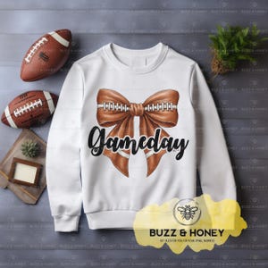 May include: White sweatshirt with a brown football-themed bow graphic and the word "Gameday" in black script. Two brown footballs are visible, along with a small wooden tray and a sprig of greenery. The sweatshirt is a casual, sporty style.