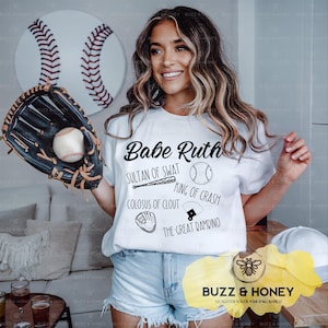 May include: A white t-shirt with the text "Babe Ruth" and baseball-related graphics. A baseball glove holds a baseball. A baseball is mounted on the wall. The shirt also has the phrases "Sultan of Swat" and "King of Crash".