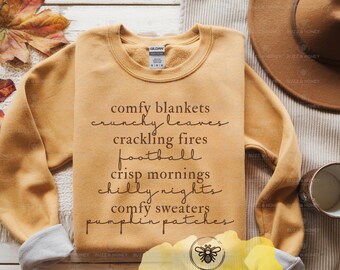May include: Mustard-colored sweatshirt with the words "comfy blankets, crunchy leaves, crackling fires, football, crisp mornings, chilly nights, comfy sweaters, pumpkin patches" in a handwritten font. A brown hat and a mug are visible.