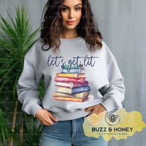 Puede incluir: Una sudadera gris con el texto "let's get lit" impreso en la parte delantera. Una pila de libros pintados con acuarela está centrada en la sudadera. La sudadera la lleva puesta una persona.