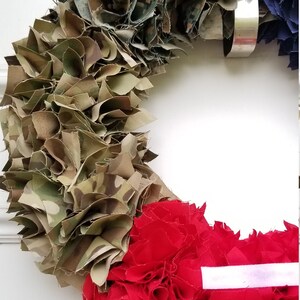 Puede incluir: Una corona patri&oacute;tica con secciones de tela de camuflaje, roja con rayas blancas y azul. La corona est&aacute; colgada en una puerta blanca. Decoraci&oacute;n.