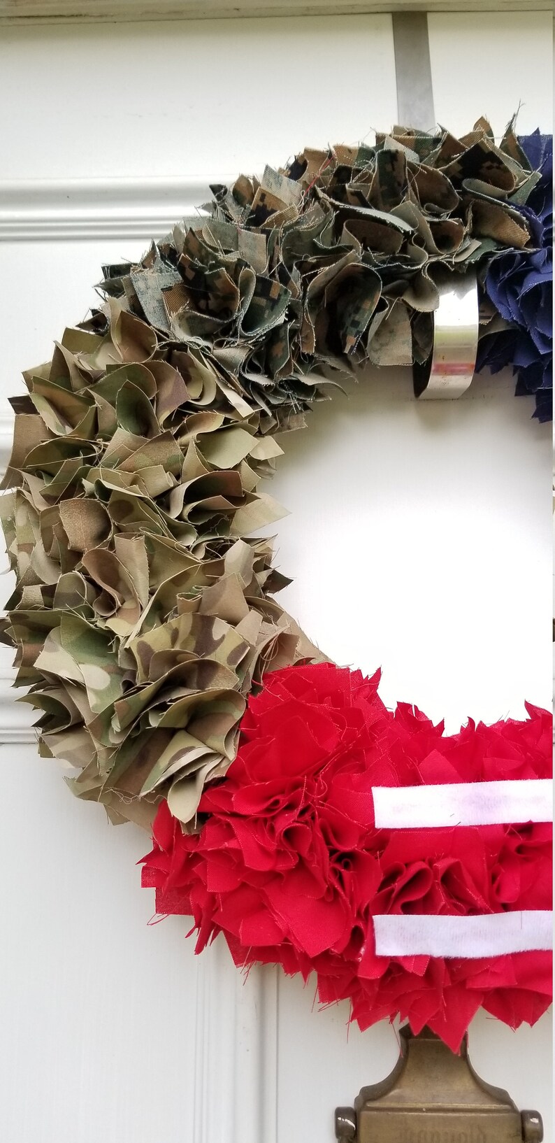 Puede incluir: Una corona patri&oacute;tica con secciones de tela de camuflaje, roja con rayas blancas y azul. La corona est&aacute; colgada en una puerta blanca. Decoraci&oacute;n.