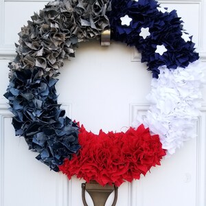 Puede incluir: Una corona circular decorativa hecha de tela gris, azul marino, blanca y roja. Estrellas blancas adornan la secci&oacute;n azul marino. La corona est&aacute; colgada en una puerta blanca con un llamador de lat&oacute;n.