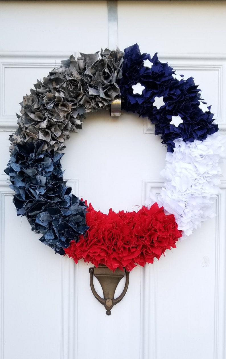 Puede incluir: Una corona circular decorativa hecha de tela gris, azul marino, blanca y roja. Estrellas blancas adornan la secci&oacute;n azul marino. La corona est&aacute; colgada en una puerta blanca con un llamador de lat&oacute;n.