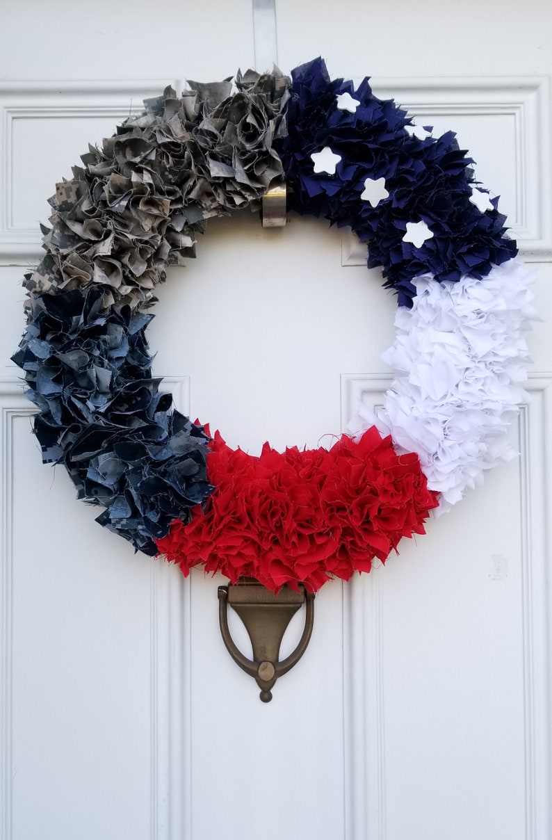 Puede incluir: Una corona patri&oacute;tica con un dise&ntilde;o circular, con secciones de tela gris, azul marino, roja y blanca. Acentos blancos en forma de estrella son visibles en la secci&oacute;n azul marino. La corona cuelga de una puerta blanca con un llamador de lat&oacute;n.