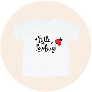 May include: White t-shirt with the words "Little Lovebug" in black script, accompanied by a red and black ladybug illustration. The shirt is a short-sleeved crew neck style, suitable for children.