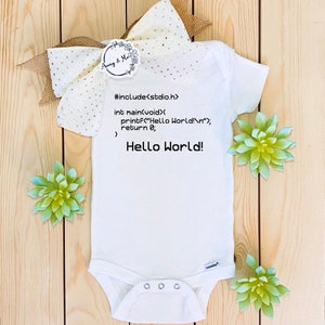 May include: White baby onesie with the text "Hello World!" and programming code. The onesie is decorated with a cream-colored bow and artificial succulents. The onesie is a newborn gift.