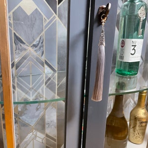 May include: A grey cabinet with glass doors and shelves. The cabinet has a gold trim and a tassel on the door handle. There are bottles of alcohol on the shelves.
