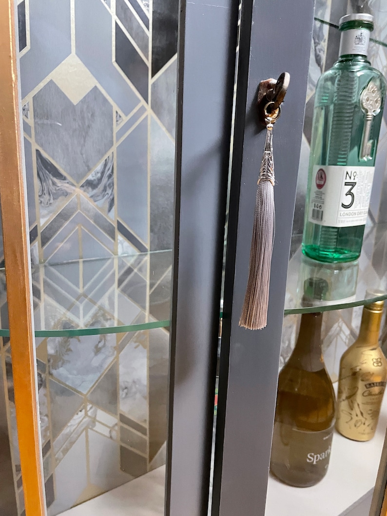 May include: A grey cabinet with glass doors and shelves. The cabinet has a gold trim and a tassel on the door handle. There are bottles of alcohol on the shelves.