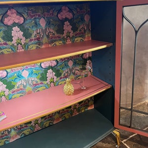 Puede incluir: Un armario azul verdoso con ribetes dorados y un interior rosa. El armario tiene dos estantes y una puerta de vidrio. Los estantes est&aacute;n forrados con papel tapiz floral. Un peque&ntilde;o pi&ntilde;a dorada y un vaso est&aacute;n en el estante superior.