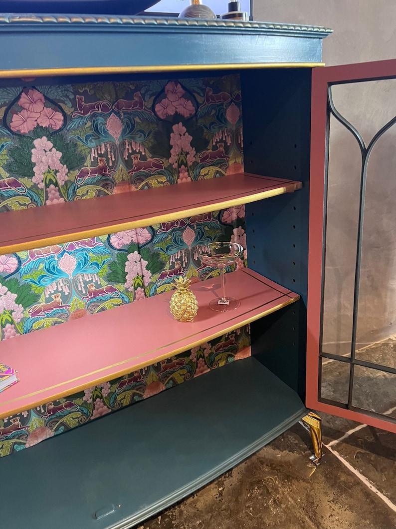 Puede incluir: Un armario azul verdoso con ribetes dorados y un interior rosa. El armario tiene dos estantes y una puerta de vidrio. Los estantes est&aacute;n forrados con papel tapiz floral. Un peque&ntilde;o pi&ntilde;a dorada y un vaso est&aacute;n en el estante superior.