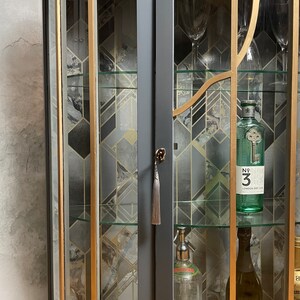 May include: A dark grey corner cabinet with glass doors and shelves. The cabinet has gold trim and a geometric pattern on the back wall. The shelves are filled with bottles of spirits and other barware.