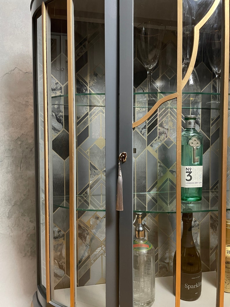 May include: A dark grey corner cabinet with glass doors and shelves. The cabinet has gold trim and a geometric pattern on the back wall. The shelves are filled with bottles of spirits and other barware.