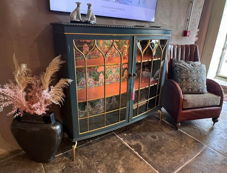 Puede incluir: Un armario verde oscuro con puertas de vidrio y detalles dorados. El armario tiene dos estantes y est&aacute; decorado con un patr&oacute;n floral. El armario est&aacute; sobre un suelo de baldosas junto a un sill&oacute;n de cuero marr&oacute;n con un coj&iacute;n estampado.