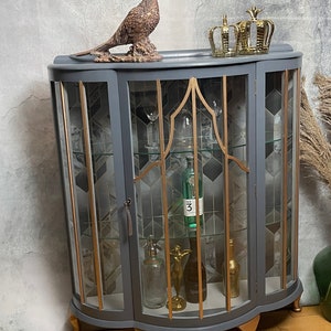 May include: A grey curved glass-fronted cabinet with gold accents and gold feet. The cabinet has three shelves and is decorated with a gold pheasant figurine and gold crowns on top.