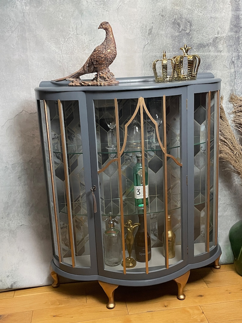 May include: A grey curved glass-fronted cabinet with gold accents and gold feet. The cabinet has three shelves and is decorated with a gold pheasant figurine and gold crowns on top.