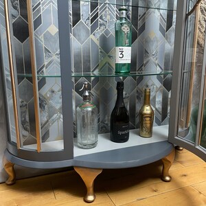May include: A grey glass-fronted cabinet with gold accents and four gold feet. The cabinet has three shelves with various bottles and glasses on display. The cabinet is on a wooden floor.