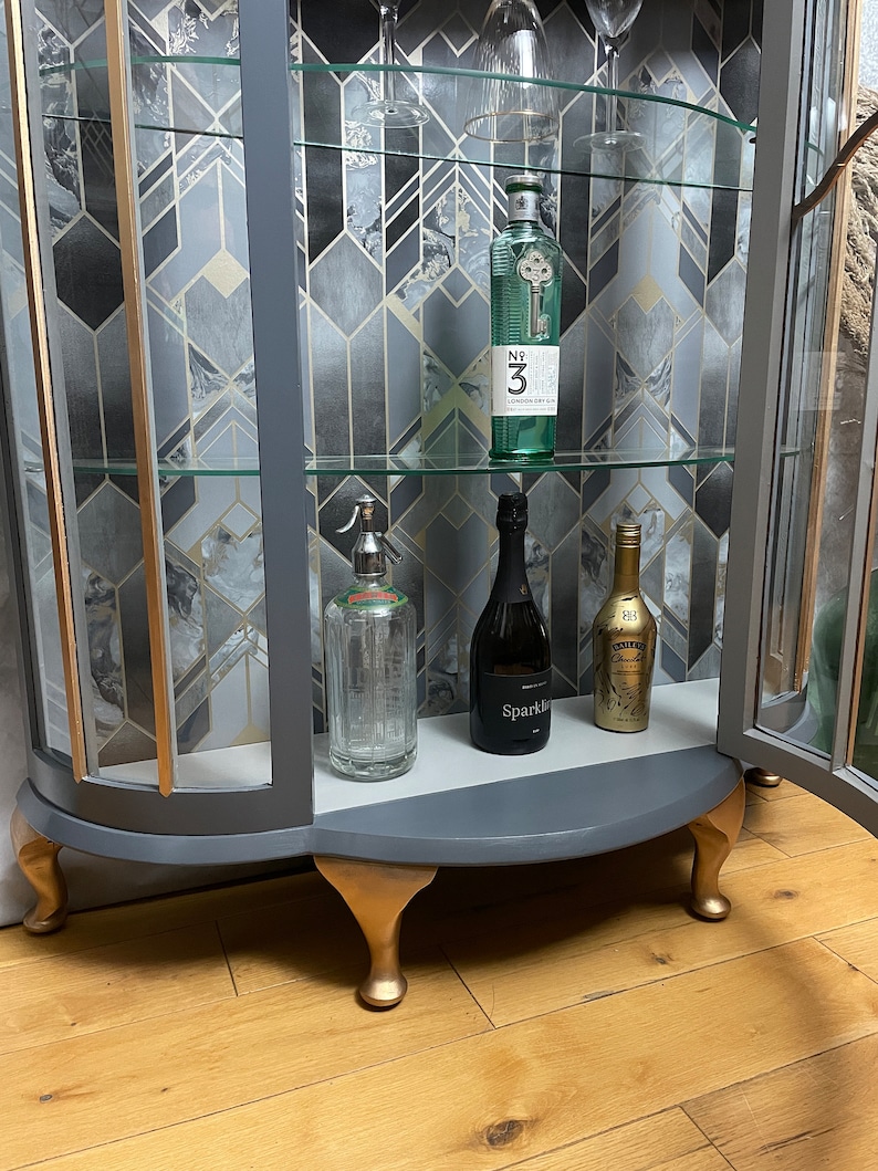 May include: A grey glass-fronted cabinet with gold accents and four gold feet. The cabinet has three shelves with various bottles and glasses on display. The cabinet is on a wooden floor.