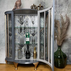 May include: A grey and gold curved glass cabinet with a glass door open. The cabinet has three shelves and is decorated with a geometric pattern. The cabinet is sitting on a wooden floor. There are bottles of alcohol and glasses on the shelves.