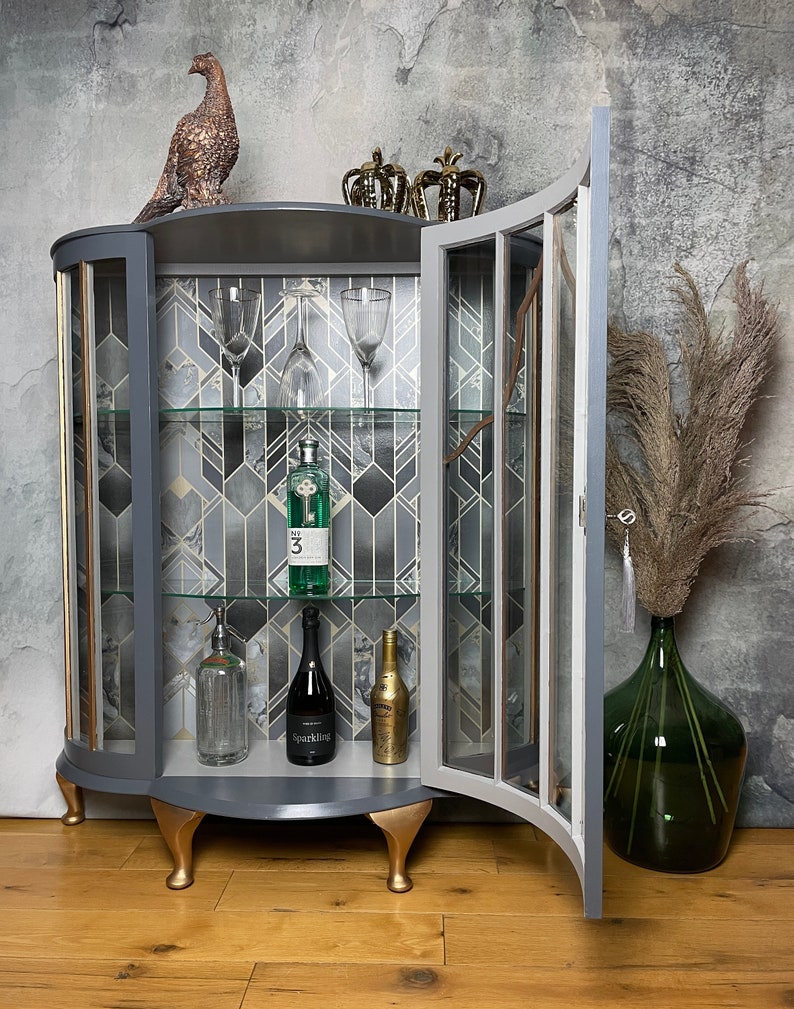 May include: A grey and gold curved glass cabinet with a glass door open. The cabinet has three shelves and is decorated with a geometric pattern. The cabinet is sitting on a wooden floor. There are bottles of alcohol and glasses on the shelves.